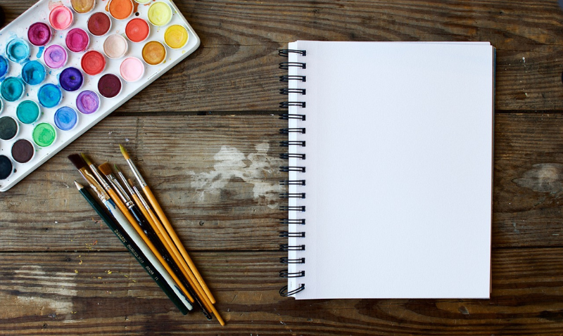 A blank spiral-bound notebook sits on a wooden surface with a set of watercolor paints and several brushes arranged next to it.