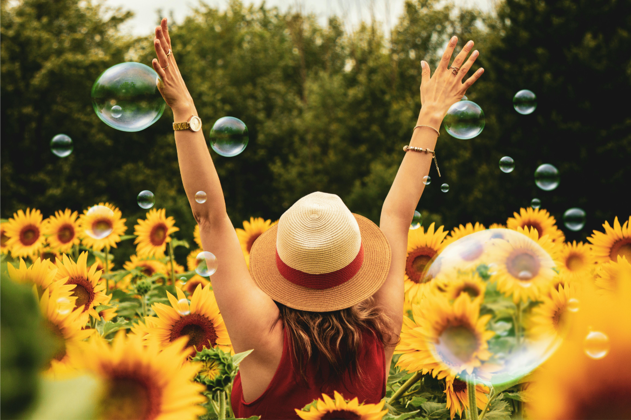 Une personne portant un chapeau de soleil et une robe rouge se tient les bras levés dans un champ de tournesols, entouré de bulles, se réjouissant de son bilan de compétences.