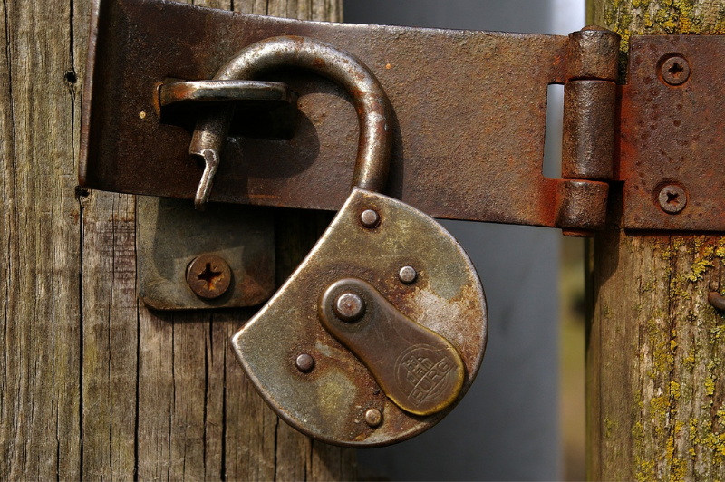 A rusty padlock with a partially inserted key hangs unlocked on an old wooden door secured with a rusty metal latch.