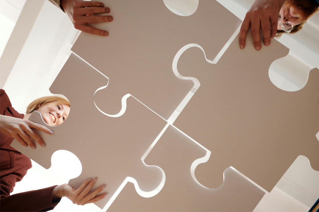 Four hands are seen connecting large white puzzle pieces. A person smiles while participating in the activity. The perspective is from below, looking at individuals and pieces of the puzzle, promoting quality of work life through collaboration and shared success.