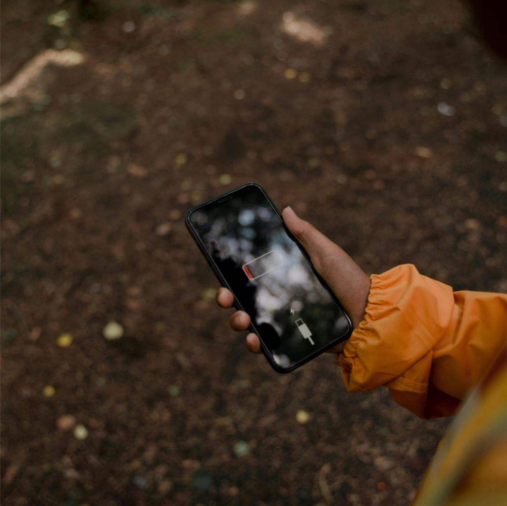 Une personne portant une veste orange tient un smartphone affichant une icône de batterie faible avec un symbole de câble de charge dans un environnement extérieur, soulignant la nécessité de faire une pause et de recharger ses propres batteries à plat.