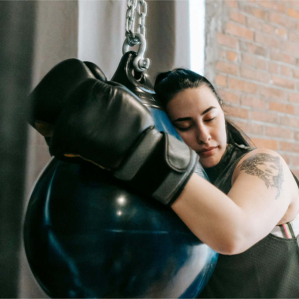 Une femme tatouée, portant des gants de boxe, pose sa tête contre un sac de boxe suspendu, incarnant la nécessité de faire une pause.