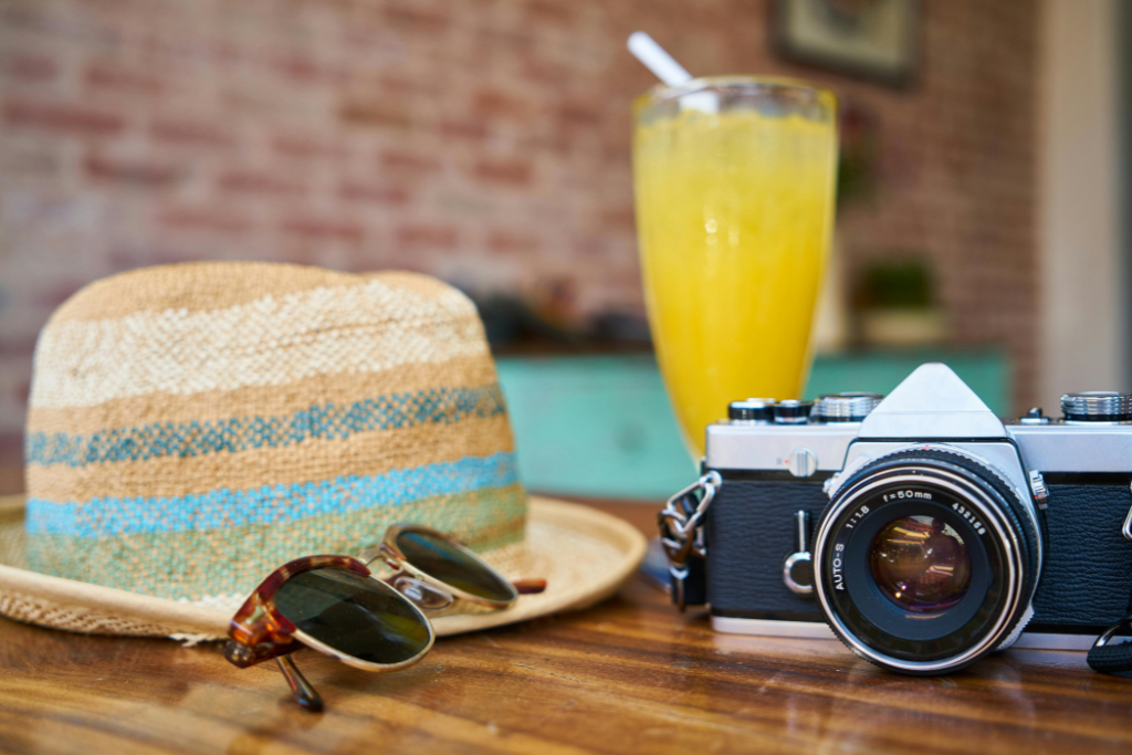 Un chapeau de paille, des lunettes de soleil, un appareil photo vintage et un verre de jus d'orange avec une paille sont placés sur une table en bois avec un mur de briques flou en arrière-plan. C'est la configuration parfaite pour faire une pause.