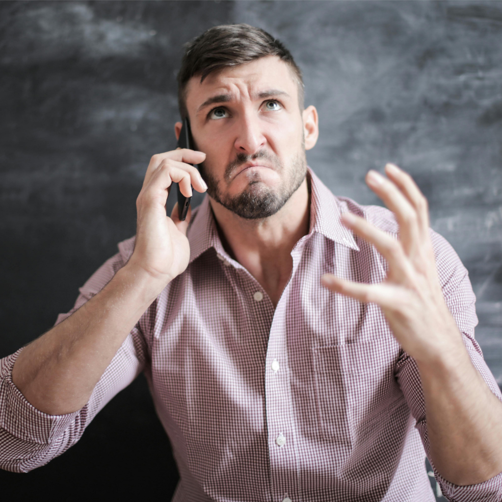 Un homme en chemise à carreaux est au téléphone, l'air frustré et levant la main gauche dans un geste exaspéré, probablement aux prises avec des conflits en équipes. Il se tient devant un fond de tableau noir.