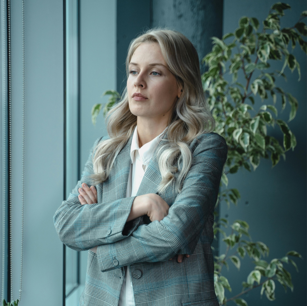 A blonde-haired woman in a checkered blazer stands with her arms crossed and looks thoughtfully out the window.