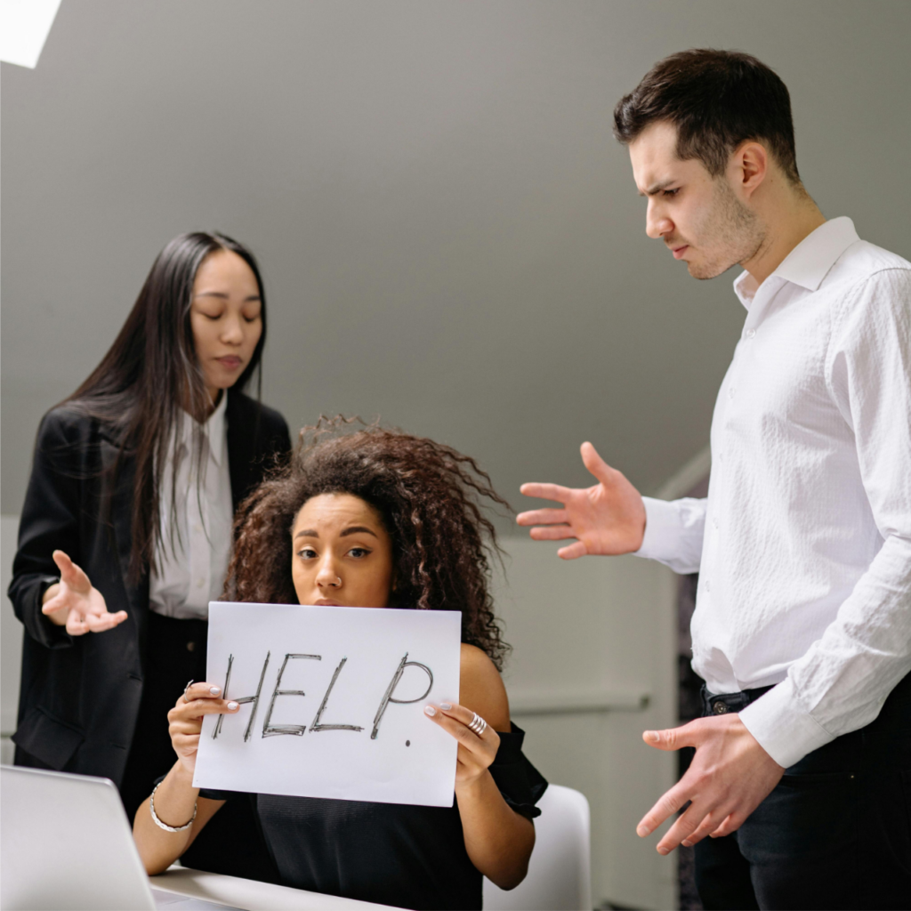 A woman holds a sign reading “HELP.” She sits at a desk with a laptop, while a man and a woman stand nearby, looking worried.