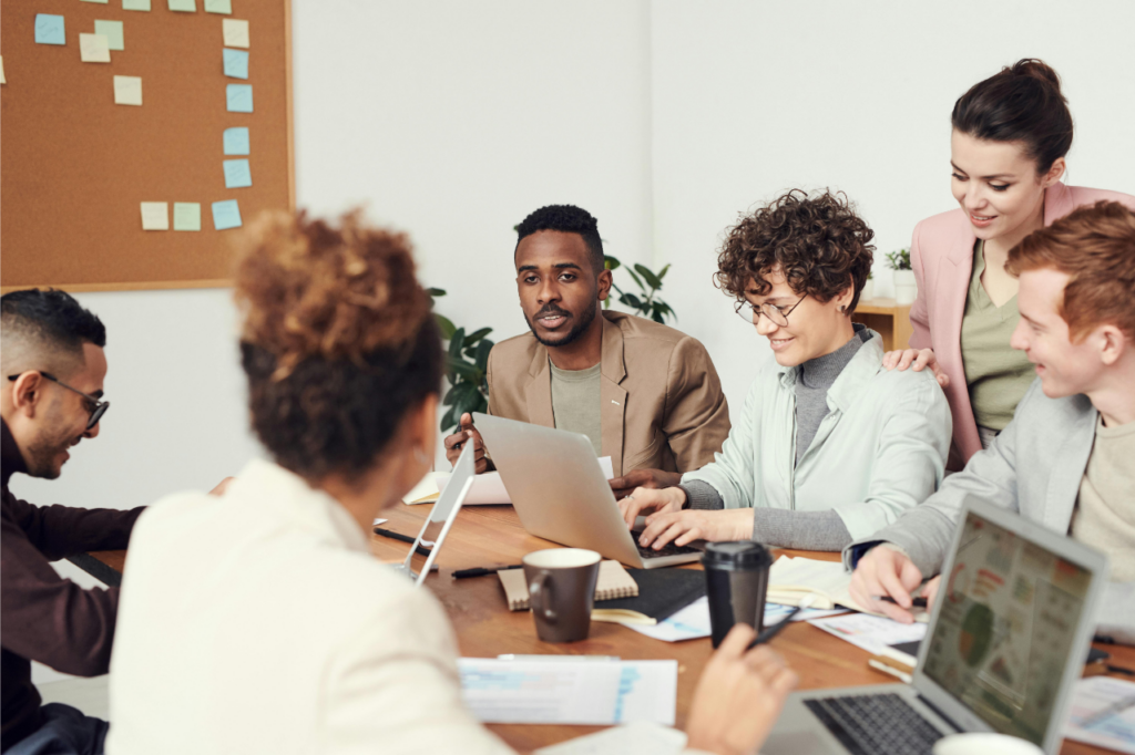 Group of six adults chatting around a table with laptops, notebooks and documents, focused on team conflict resolution. Cork board with sticky notes in the background.