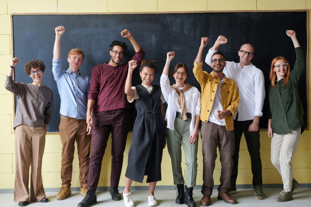 A diverse group of eight adults stand in front of a blackboard, smiling and raising their right fists, united in their commitment to promoting mental health in business.