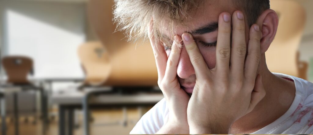 A person with closed eyes holds their face in their hands, looking stressed or scared. The blurred background of chairs and tables adds to the feeling of isolation.