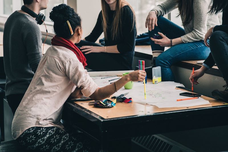 A group of people sit around a table with papers and markers, engaging in lively discussion while contributing to a mental health mural.