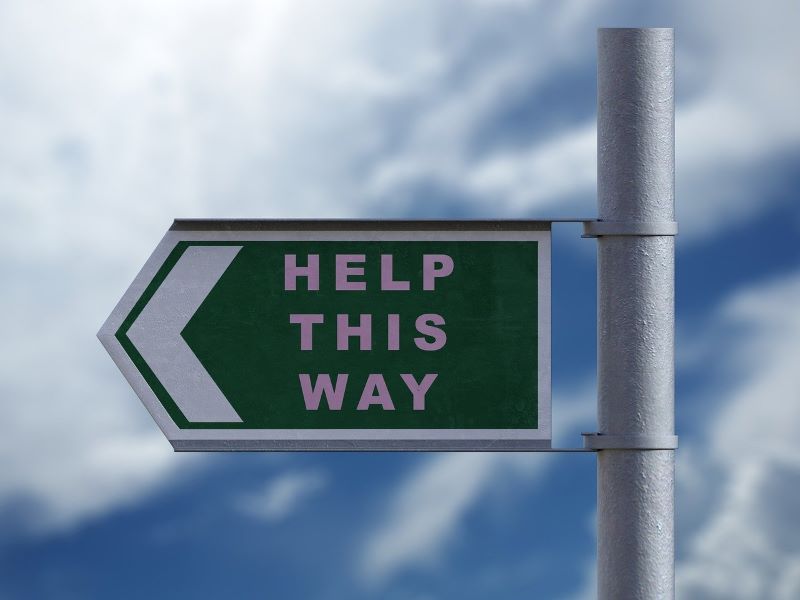 A directional sign with the message “Help This Way” stands against a backdrop of cloudy skies, reminiscent of a mural depicting mental health pathways.