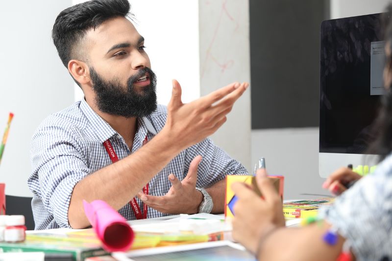 A bearded man gestures while discussing a mental health mural with a colleague seated at a desk, the computer and colorful stationery adding vibrancy to their collaborative environment.