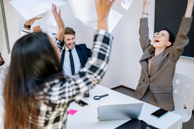 In an office buzzing with energy, a group of people celebrate triumphantly, throwing papers into the air as if to pick up the pace after the festivities. A laptop and notepads sit on the table, evidence of their renewed vigor and enthusiasm.