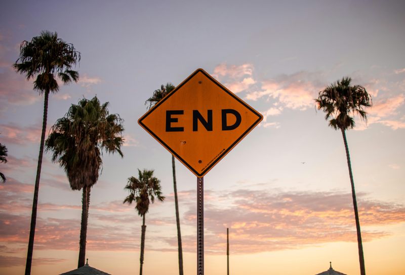 A sign reading “END” stands in front of tall palm trees, set against a tranquil sunset sky, as if to encourage people to pick up the pace after the festivities.