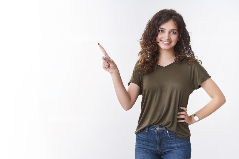 Une personne souriante aux cheveux longs et bouclés, vêtue d'une chemise verte et d'un jean, pointe énergiquement un doigt sur le côté, prête à reprendre le rythme après les fêtes.