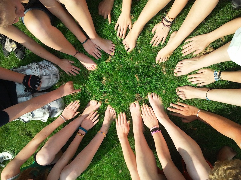 Un cercle de personnes assises sur l'herbe avec les mains et les pieds tendus vers le centre, favorisant la santé mentale en entreprise par la connexion et la pleine conscience.