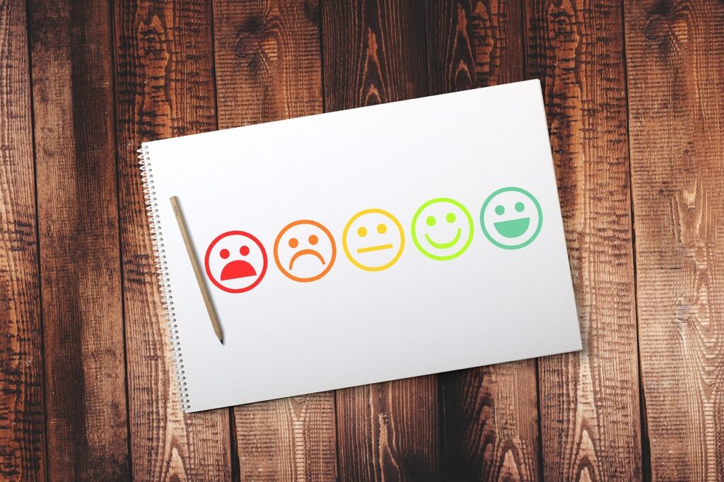 A Corporate Mental Health First Aid notebook features six smiling faces ranging from sad red to happy green, next to a pencil placed on a wooden surface.