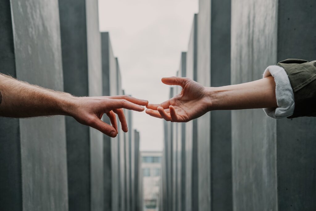 Two hands outstretched towards each other, against a background of tall, narrow and rectangular structures, symbolizing the bond and support found in First Aid for Mental Health in Business.